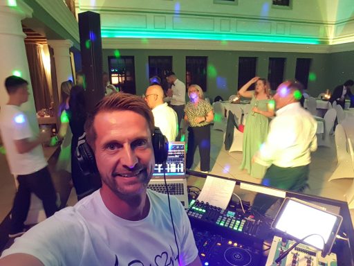 DJ beim Auflegen auf einer Party, mit tanzenden Gästen und buntem Licht im Hintergrund.